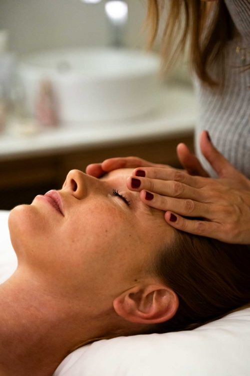 Mujer rubia recibiendo tratamiento facial. La especialista tiene sus manos apoyadas sobre la frente de la clienta. En el fondo de la foto se ve un lavabo desenfocado