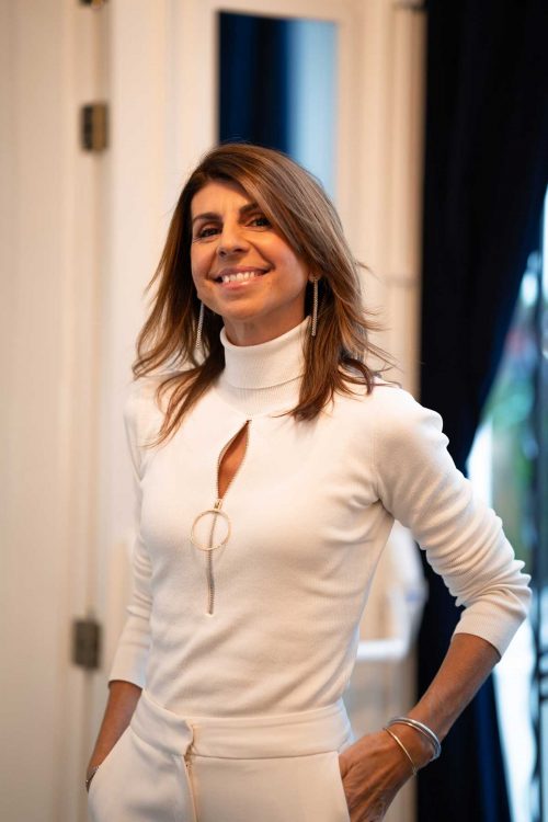 Retrato de Maria Casado creadora del metodo Maricasado, especialista en pieles y tratamiento faciales de autor. lleva un jersey blanco de cuello vuelto y un pantalón blanco ajustado. Esta con una postura relajada y una mano en el bolsillo. Esta sonriente y Feliz